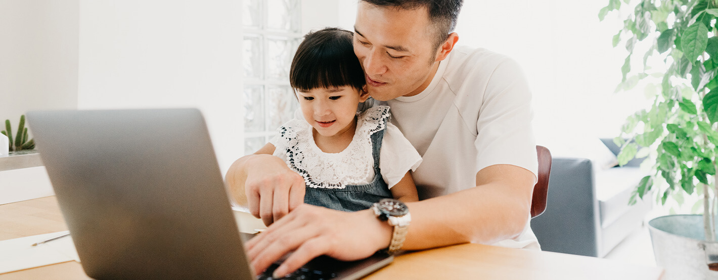 A father sits at a table with his daughter in his lap while researching how to save for college on his laptop.