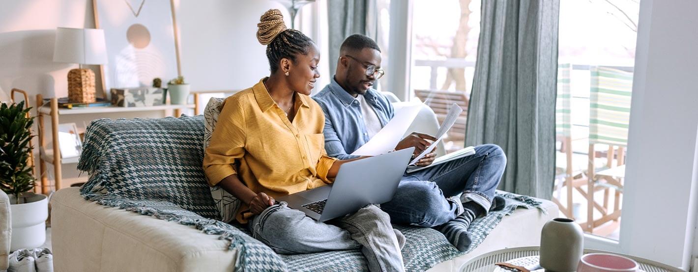 A man and a woman sit on the couch together reviewing documents while looking for an online budget calculator.